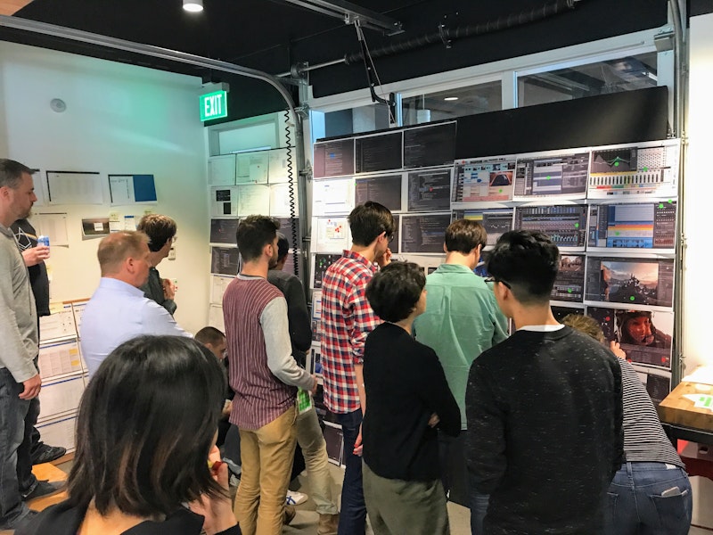 A group of designers is observing and discussing screenshots printed on numerous papers stuck to the wall.